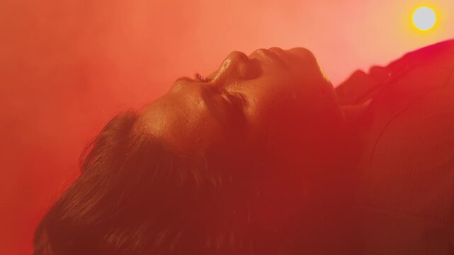 Close-up profile of a woman with glistening skin lying down in an orange, smoky environment. Conveys a sense of intense heat, fever, or extreme exhaustion.