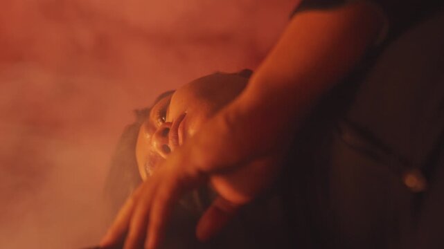 A low-angle, stylized shot of a woman with closed eyes, gently resting her hand on her body in a red, smoky room. Conveys self-love, vulnerability, and deep relaxation.