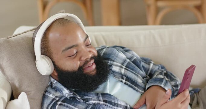 Vertical video: Relaxing man in plaid opening eyes, scrolling phone with headphones on sofa at home