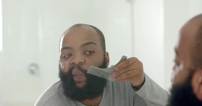 Man checking bathroom mirror, holding wide comb, combing beard and noting scissors for symmetry