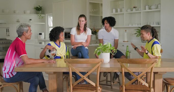 Diverse friends sitting at wood table in kitchen chatting around soccer placing ball drinking beer