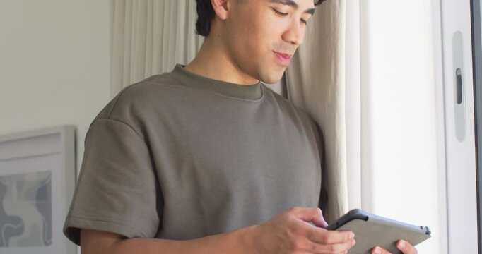 Man standing at window holding tablet, tapping scrolling and reading in olive tee