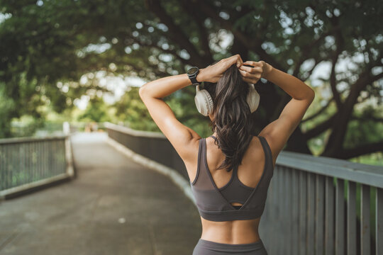 Asian fit woman tying hair back in park after workout. Rear view. Active lifestyle, fitness, wellness, and mental health concept. Wearing sportswear, headphones, and smartwatch.