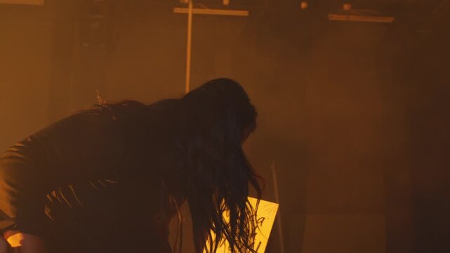 An emotional woman bends down in despair on a foggy stage bathed in warm orange light, as faceless figures hold up insulting signboards. A powerful metaphor for online hate and bullying.