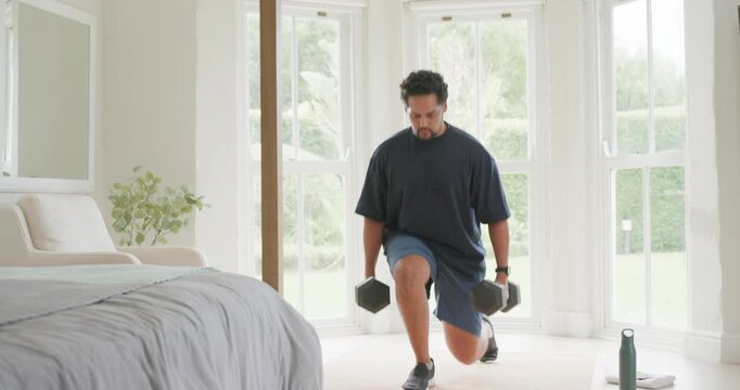 African American man in sportswear standing in bedroom, doing lunges with hex dumbbells for fitness