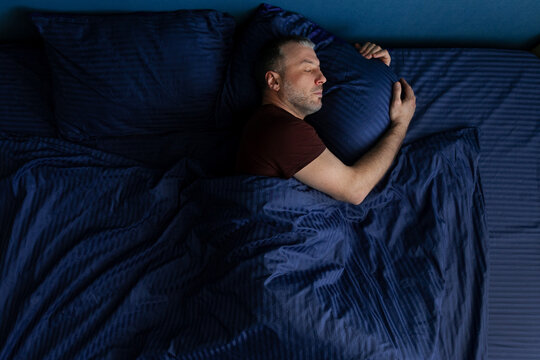 Top view of middle aged man sleeping on side hugging pillow tightly