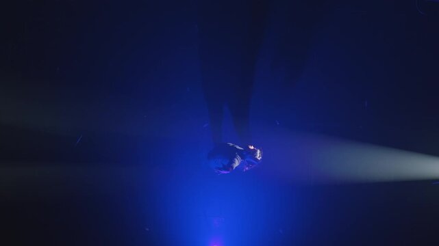 A cinematic overhead view of a female dancer lying on a dark stage, spreading her arms wide under a moody blue spotlight. The pose suggests surrender, exhaustion, or emotional release.