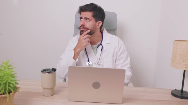 Young pensive male doctor in a white coat with a stethoscope sitting at his office desk with a laptop, thoughtfully considering a patient's diagnosis or a complex medical problem