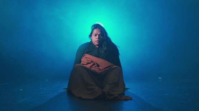 A female performer suddenly sits up from the floor, looking shocked and bewildered in a cinematic blue spotlight. Represents waking from a nightmare or resurrection.