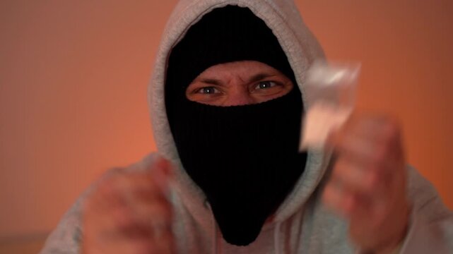 Close-up of a narcotic substance in a transparent bag held by a man in a black mask against the backdrop of police arriving and flashing lights, drug trafficking concept