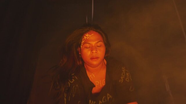 A troubled young woman with abusive words written on her face and body stands in thick orange smoke, holding a vintage telephone receiver to her ear in shock.