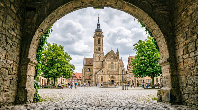 Vista de una iglesia hist&oacute;rica enmarcada por un arco de piedra. La plaza empedrada y los &aacute;rboles verdes destacan bajo un cielo nublado, creando una atm&oacute;sfera de serenidad y patrimonio antiguo.