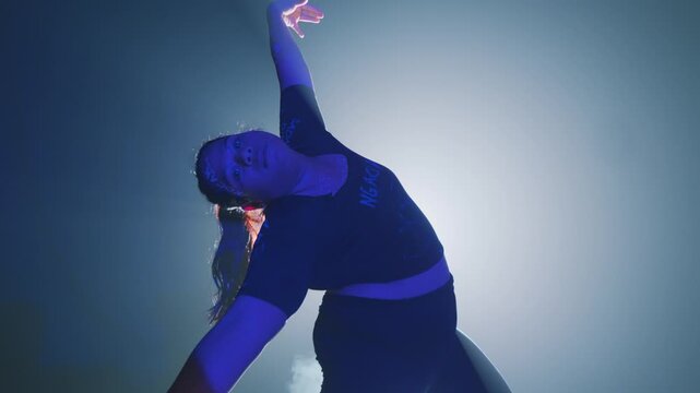A strong woman bearing painted insults on her skin performs a vigorous, emotional dance in a foggy studio. Her fierce expressions and dynamic moves symbolize the active fight against body shaming.