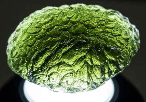 Detailed macro photograph of a vibrant, translucent green moldavite tektite, showcasing its unique, bubbly, and textured surface with a soft light illuminating from below.