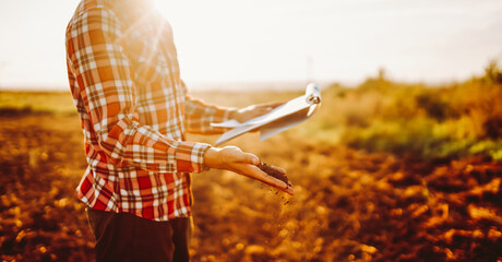 Young farmer in checkered shirt holds fertile soil and tablet in a field. Agronomist with tablet checks soil quality in the sun's rays. Concepts of agriculture, gardening, and ecology. © maxbelchenko