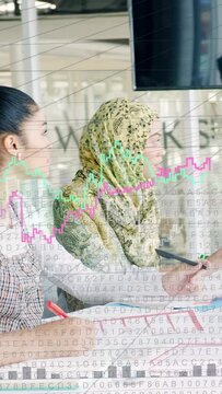 Two women sitting at table, business monitor showing graphs and data, one pointing, other noting