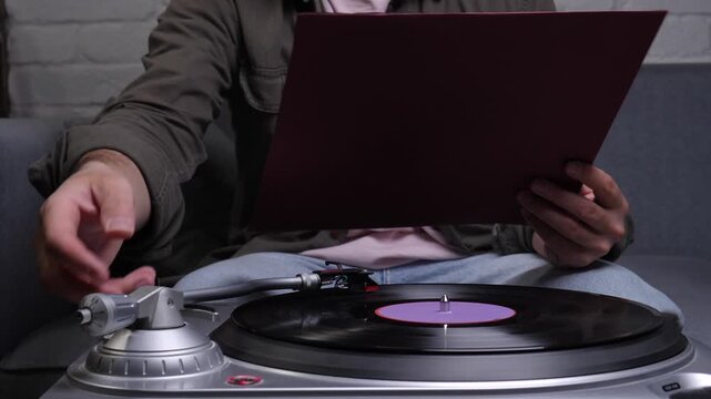 Person lounges on a sofa at home, holding an album cover while a vinyl record spins on a turntable, enjoying relaxed retro audio nostalgia and cozy evening listening time