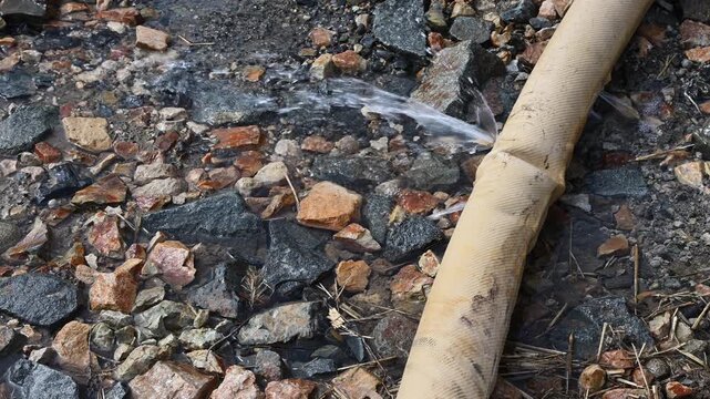 Large industrial tan hose with a hole and water leaking, spraying under pressure, out onto ground at construction site
