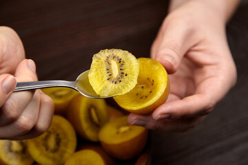 Obraz premium Hands holding a spoon with a slice of golden kiwi fruit above a halved kiwi on a wooden table with additional whole kiwis in the background