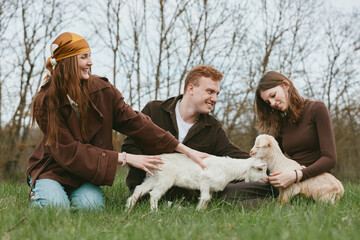 Fototapeta premium A joyful trio outdoors with a playful goat kid, as friends gently pat the animals. Casual lifestyle scene highlighting bonding, care, and rural simplicity.