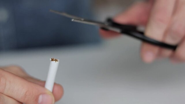 Close-up of a hand holding a cigarette while scissors cut it in half, symbolizing quitting smoking, bad habit rejection and healthy lifestyle concept.