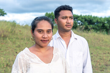 Obraz premium A cinematic portrait of a young Indonesian couple in white clothing, posing together in a lush meadow with a dramatic mountain ridge background. 