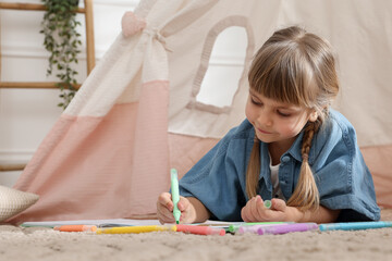 Little girl drawing in sketchbook near toy wigwam at home © New Africa