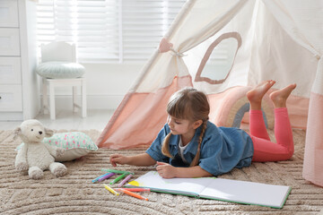 Little girl drawing in sketchbook near toy wigwam at home © New Africa