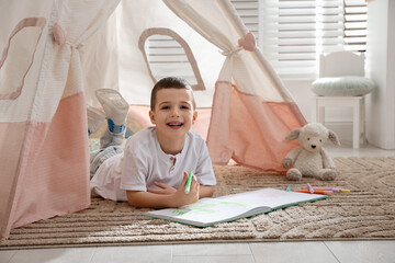 Little boy drawing in sketchbook near toy wigwam at home © New Africa