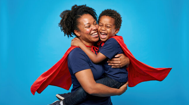 African American mother lifting her laughing young son while wearing red superhero capes against a vibrant blue studio background, celebrating supermom and family bond.