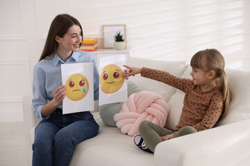 Professional psychologist showing pictures of sad and happy faces to little girl on sofa indoors