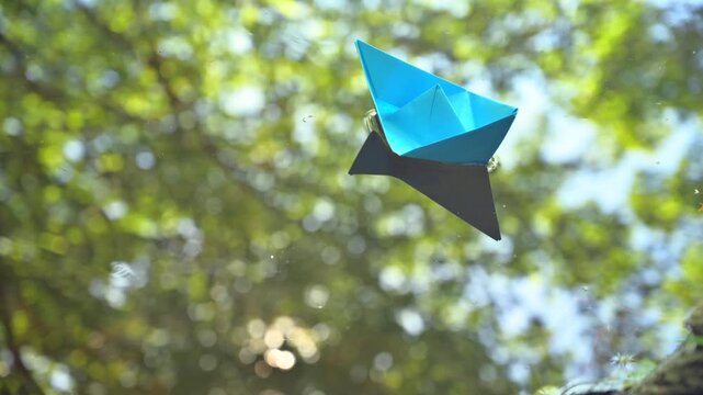 Conceptual video of a small blue paper boat slowly drifting on calm water, reflecting a rich canopy of leafy trees. Peaceful and poetic atmosphere representing nature, childhood imagination, serenity 