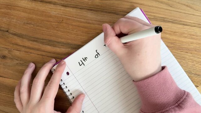 Close up of hands writing &ldquo;4th of July plans&rdquo; on lined notebook paper, summer holiday planning concept for Independence Day, natural light overhead shot on wooden table, 4K video