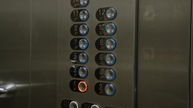 Hand presses illuminated elevator button on metal control panel, closeup of numbered buttons and glowing indicator, tactile