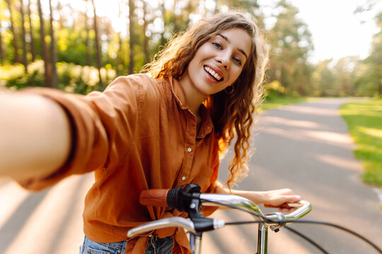 A cheerful woman takes a selfie on her phone while riding a bicycle in a sunny park. A young woman enjoys the sunset and rides a bicycle outdoors. Concept: relaxation, weekends, and blogging.