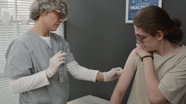 Dolly out shot of Caucasian teenager getting vaccinated in modern hospital, mature Caucasian nurse placing band aid onto his shoulder, perfect for immunization campaigns