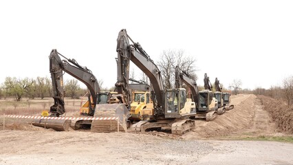 Rząd nowoczesnych koparek gąsienicowych stoi na placu budowy. Potężne, żółte maszyny z długimi ramionami są gotowe do robót ziemnych. W tle widać drzewa i błękitne niebo z delikatnymi chmurami © tomo81