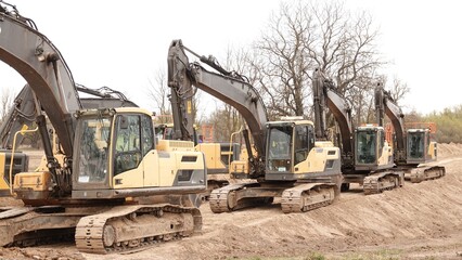 Rząd nowoczesnych koparek gąsienicowych stoi na placu budowy. Potężne, żółte maszyny z długimi ramionami są gotowe do robót ziemnych. W tle widać drzewa i błękitne niebo z delikatnymi chmurami © tomo81