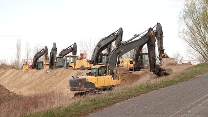 Rząd nowoczesnych koparek gąsienicowych stoi na placu budowy. Potężne, żółte maszyny z długimi ramionami są gotowe do robót ziemnych. W tle widać drzewa i błękitne niebo z delikatnymi chmurami © tomo81