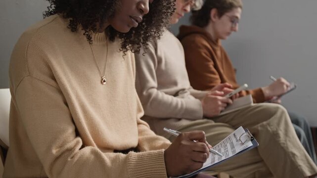 Group of three diverse patients sitting on chairs in waiting room in modern clinic, filling in paper forms and using phone