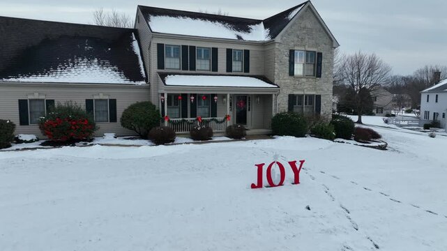 Aerial view of American suburban home with snow-covered yard and porch decorated for Christmas, featuring &ldquo;JOY&rdquo; sign. Festive winter daytime residential atmosphere.