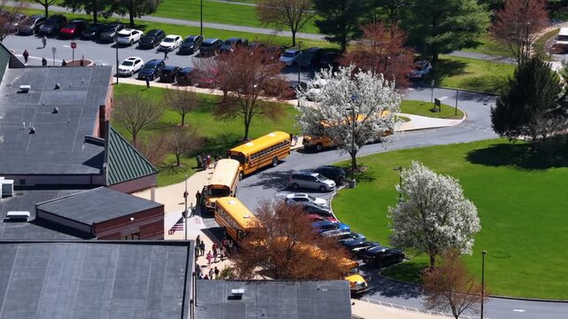 Aerial view of school campus in spring with yellow school buses picking up students, blooming trees and green lawns under bright sunlight in quiet American suburban setting. Orbit shot.