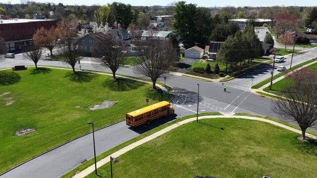 Aerial view of classic yellow school bus driving through suburban intersection in USA during spring, with green lawns, quiet streets and bright sunlight in calm neighborhood setting. Tracking shot.