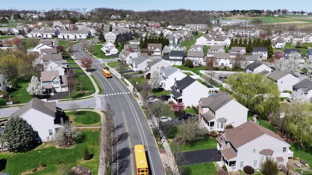 U.S. suburban neighborhood in spring with two-story homes with porches, many yellow school bus on residential street and blooming trees in quiet community. Aerial rising wide shot.