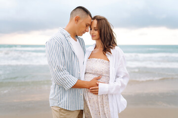 Obraz premium A pregnant woman and her man stand on the seashore, touching each other. A young married couple enjoys the seascape and relaxes outdoors. Concepts of family, love, and fusion.