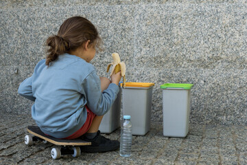 Naklejka premium Back view of a child sitting on a skateboard and eating a banana in front of three recycling bins, representing a break in an outdoor classroom.