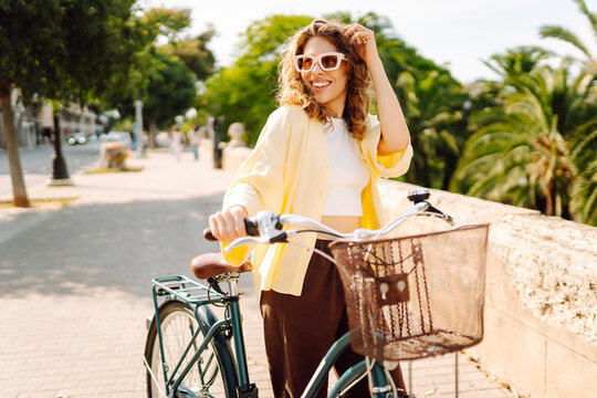 A woman rides her bicycle down a path in a city. Palm trees line the street, and she smiles while wearing sunglasses. The sun shines brightly on a clear day. Lifestyle.