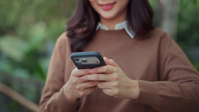 Asian woman using smartphone in city park. Close up hands holding mobile phone for social media, digital banking, and communication. Happy girl outdoor with technology.
