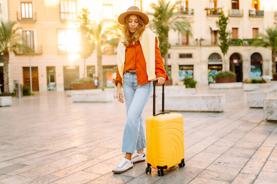 Young traveler wearing hat and carrying small suitcase stands in sunny square. Woman with backpack admires architecture while strolling through city. Concepts for travel, tourism, weekends.