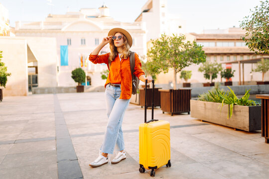 Young traveler wearing hat and carrying small suitcase stands in sunny square. Woman with backpack admires architecture while strolling through city. Concepts for travel, tourism, weekends.